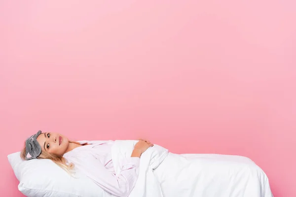 Mujer joven en pijama y con los ojos vendados mirando a la cámara mientras está acostada sobre ropa de cama sobre fondo rosa - foto de stock
