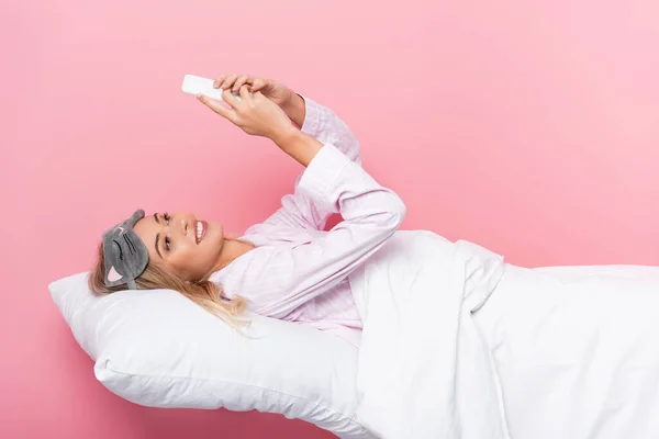 Donna sorridente in benda utilizzando smartphone su biancheria da letto su sfondo rosa — Foto stock