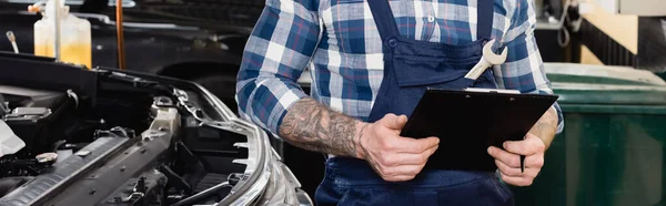 Cropped view of mechanic holding clipboard near car engine compartment, banner — Stock Photo