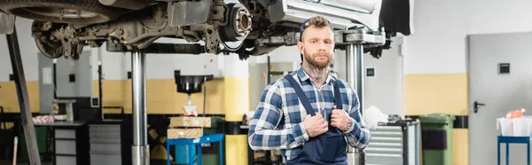 Giovane tecnico toccando cinghie di tuta mentre in piedi vicino auto sollevato su ascensore, banner — Foto stock