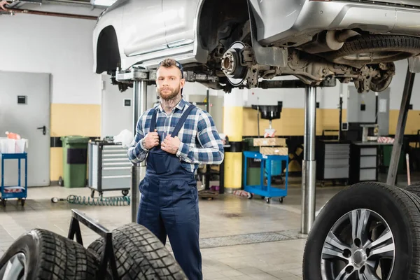 Repairman touching straps of overalls near wheels and automobile raised on car lift — Stock Photo