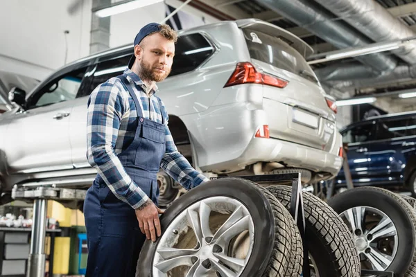 Young mechanic looking at camera near wheel and auto raised on car lift — Stock Photo