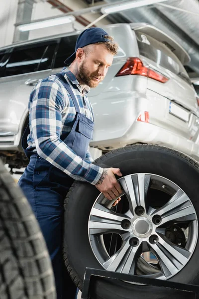 Joven técnico sosteniendo rueda cerca de coche levantado en primer plano borrosa - foto de stock