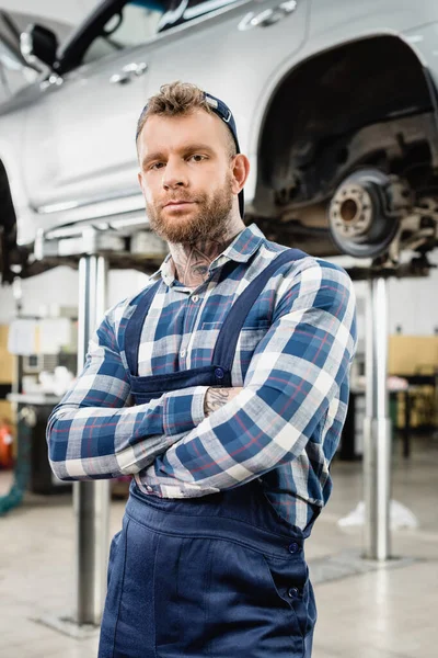 Joven reparador mirando a la cámara mientras está de pie con los brazos cruzados cerca del coche levantado sobre fondo borroso - foto de stock