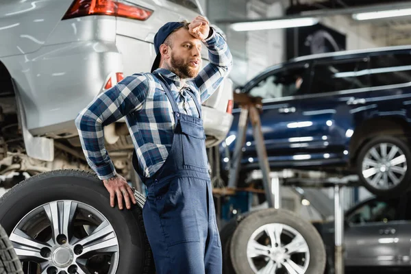 Stanco tecnico che tiene la mano vicino alla fronte mentre appoggiato sulla ruota dell'auto su sfondo sfocato — Foto stock