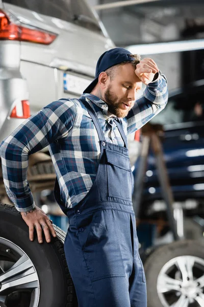 Exhausted mechanic holding hand near forehead while standing near car wheel with closed eyes on blurred background — Stock Photo