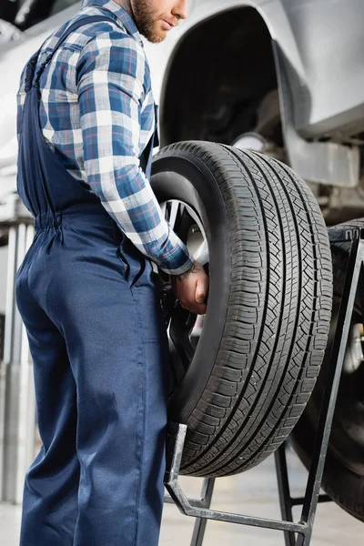 Ausgeschnittene Ansicht eines Mechanikers, der das Lenkrad neben angehobenem Auto hält, auf verschwommenem Hintergrund — Stockfoto