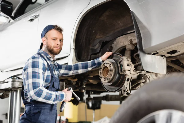 Techniker blickt auf Kamera, während er neben angehobenem Auto mit Schraubenschlüssel im verschwommenen Vordergrund steht — Stockfoto