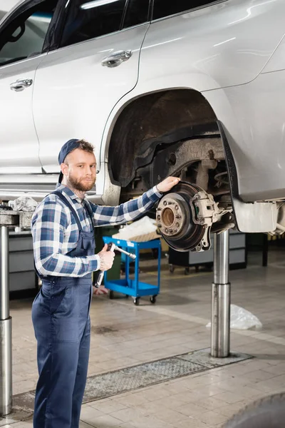 Young mechanic looking at camera while standing near auto, raised on car lift — Stock Photo