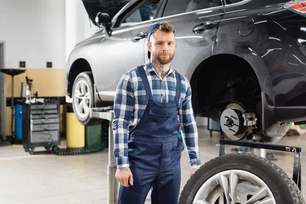 Mechanic in overalls looking at camera near automobile raised on car lift — Stock Photo