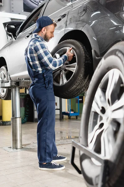 Technician in overalls fixing rear wheel on raised car on blurred foreground — Stock Photo