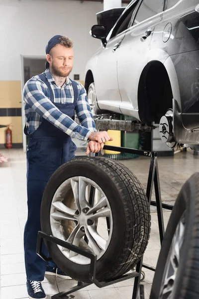 Techniker in Arbeitskleidung in der Nähe von Rad und Auto angehoben auf Autolift auf unscharfen Vordergrund — Stockfoto