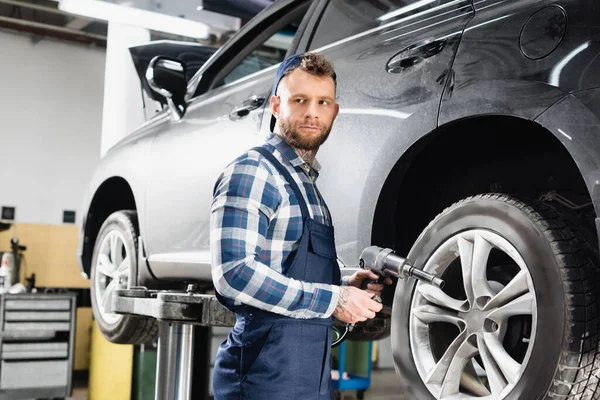 Junger Techniker schaut weg, während er Druckluftschlüssel in der Nähe des Rades des angehobenen Autos hält — Stockfoto