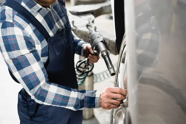 Partial view of mechanic in overalls adjusting car wheel with pneumatic wrench on blurred foreground — Stock Photo