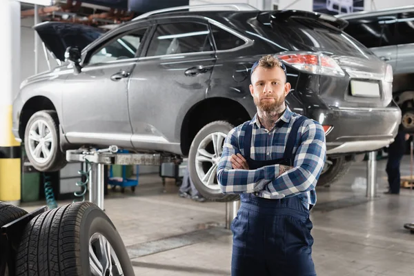 Mechanic in overalls standing with crossed arms near wheels and lifted car — Stock Photo