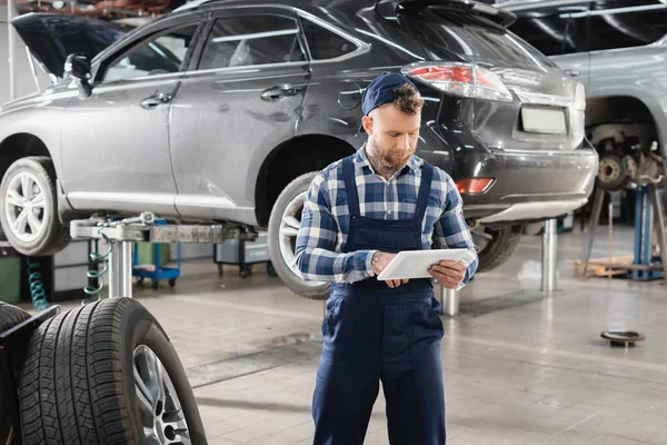 Technicien en uniforme utilisant tablette numérique près du véhicule soulevé sur ascenseur de voiture — Photo de stock