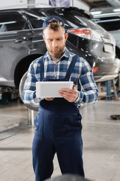 Young mechanic in overalls using digital tablet while standing near lifted car — Stock Photo