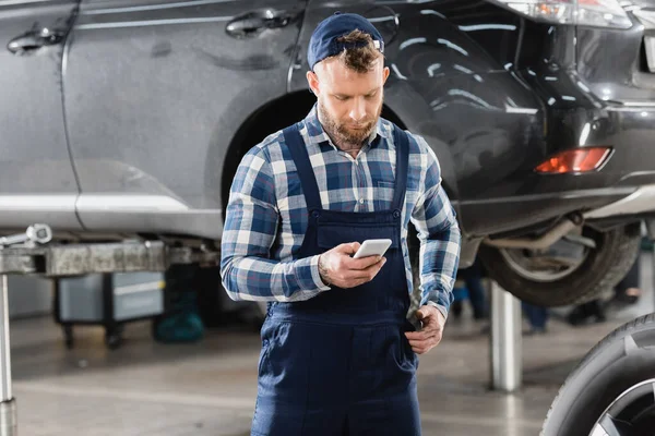 Mechanic in overalls messaging on smartphone while standing near raised car — Stock Photo