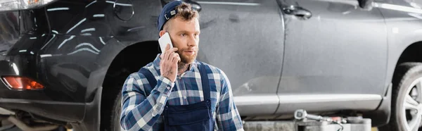 Молодой ремонтник разговаривает по мобильному телефону в мастерской возле автомобиля, баннер — стоковое фото