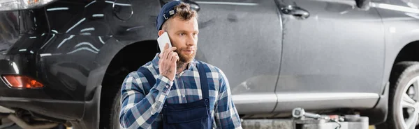 Jovem reparador falando no telefone celular em oficina perto do carro, banner — Fotografia de Stock