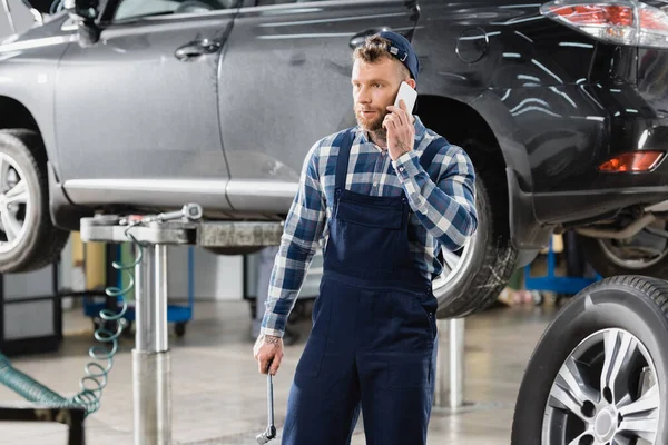 Reparador segurando chave inglesa e falando no telefone celular perto do carro levantado — Fotografia de Stock
