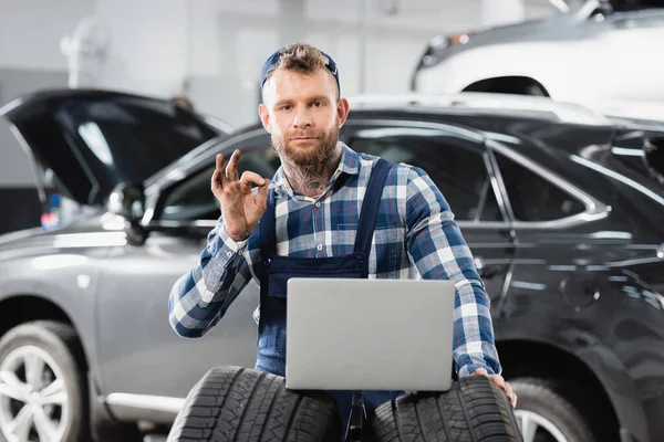 Technician showing okay gesture while holding laptop near cars on blurred background — Stock Photo