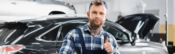 Giovane meccanico che mostra il pollice verso l'alto mentre guarda la fotocamera in officina, banner — Foto stock