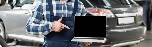 Частичное представление механика, указывающего на ноутбук с чистым экраном рядом с автомобилем на размытом фоне, баннер — стоковое фото
