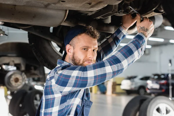 Joven mecánico mirando a la cámara mientras revisa la parte inferior del coche levantado - foto de stock