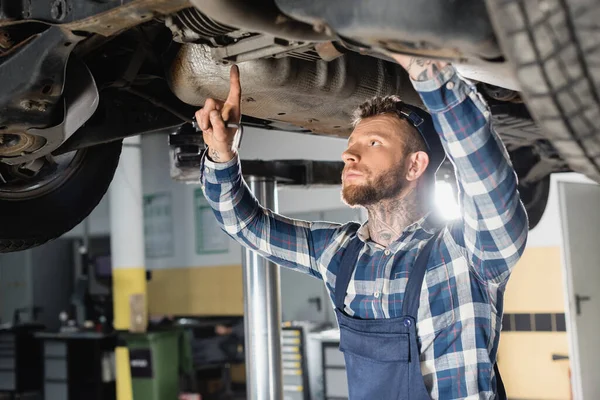 Joven reparador examinando parte inferior del automóvil levantado en el taller - foto de stock