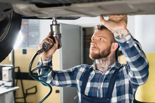 Technician repairing bottom of lifted auto with pneumatic wrench on blurred foreground — Stock Photo