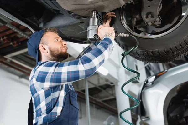 Giovane tecnico riparazione fondo di auto sollevata con chiave pneumatica — Foto stock