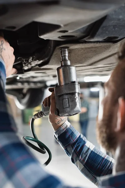 Cropped view of mechanic fixing detail of raised car with pneumatic wrench on blurred foreground — Stock Photo