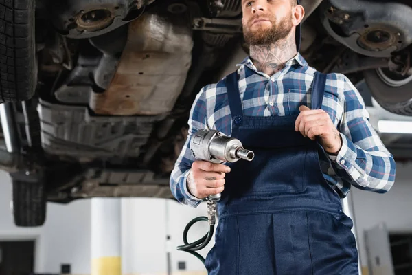 Niedrigwinkel-Ansicht des Technikers, der einen Druckluftschlüssel in der Nähe eines angehobenen Autos auf verschwommenem Hintergrund hält — Stockfoto