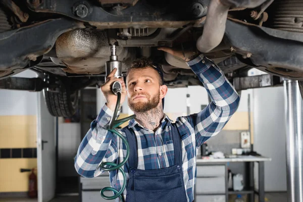 Mechanic fixing detail with pneumatic wrench while standing under raised car — Stock Photo