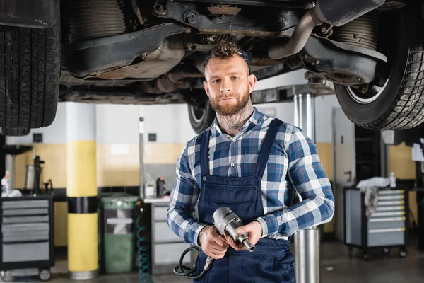Technician holding pneumatic wrench while standing under lifted car — Stock Photo