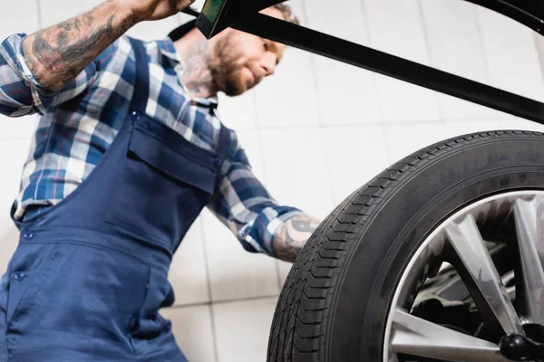 Tattooed mechanic examining wheel on balance control machine in workshop on blurred background — Stock Photo