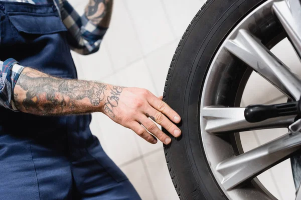 Partial view of tattooed technician touching car wheel in workshop on blurred background — Stock Photo