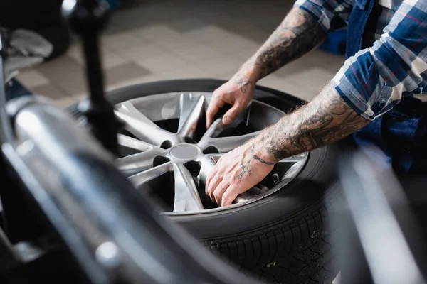 Visão parcial do reparador tatuado tomando roda de carro em oficina em primeiro plano desfocado — Fotografia de Stock