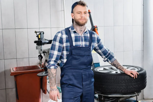 Giovane meccanico in tuta da lavoro in piedi vicino alla ruota sulla macchina di sostituzione pneumatici in officina — Foto stock