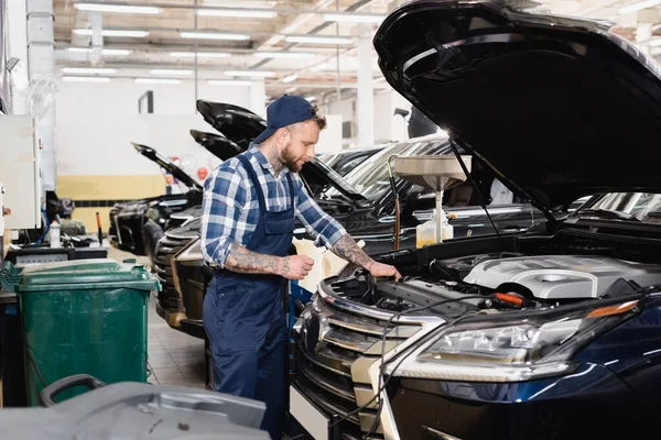 Jeune mécanicien tenant chiffon tout en se tenant près de la voiture avec capot ouvert dans l'atelier — Photo de stock