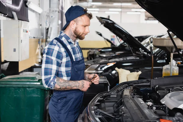 Junger Mechaniker in Arbeitskleidung steht neben Auto mit geöffneter Motorhaube in Werkstatt — Stockfoto