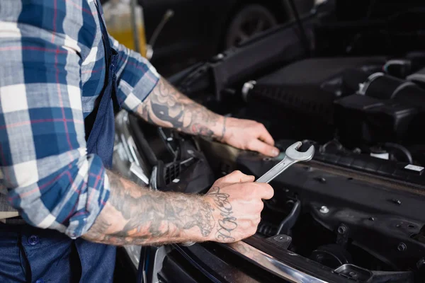 Vista parcial del técnico tatuado que sostiene la llave inglesa cerca del compartimiento del motor del coche en el primer plano borroso - foto de stock