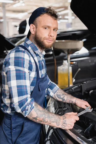 Mecánico tatuado que sostiene la llave mientras está parado cerca del compartimiento del motor del coche - foto de stock