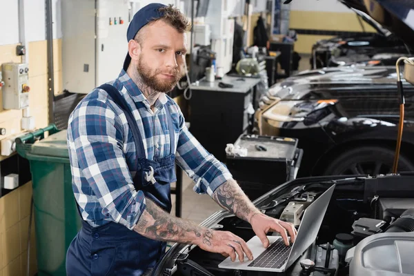 Mechanic looking at camera while making car diagnostic on laptop near engine compartment — Stock Photo