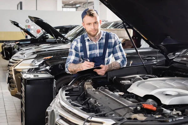 Repairmen holding clipboard and looking at camera near car engine compartment — Stock Photo