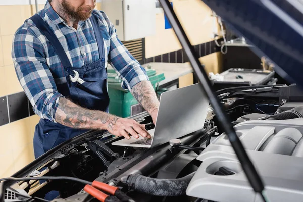 Partial view of tattooed repairman making car engine compartment diagnostic with laptop on blurred foreground — Stock Photo