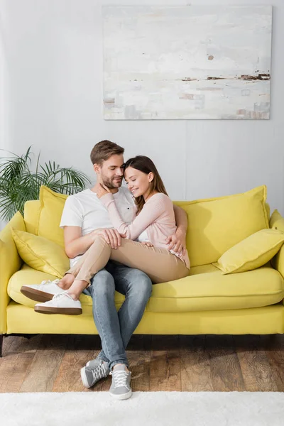 Fröhliche Mann und Frau umarmen sich, während sie auf dem Sofa im Wohnzimmer sitzen — Stockfoto