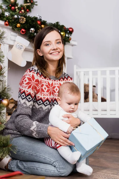 Femme gaie tenant présent près de bébé garçon dans appartement décoré — Photo de stock