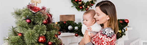 Lächelnde Frau mit Baby im Arm in der Nähe des geschmückten Weihnachtsbaums, Banner — Stockfoto